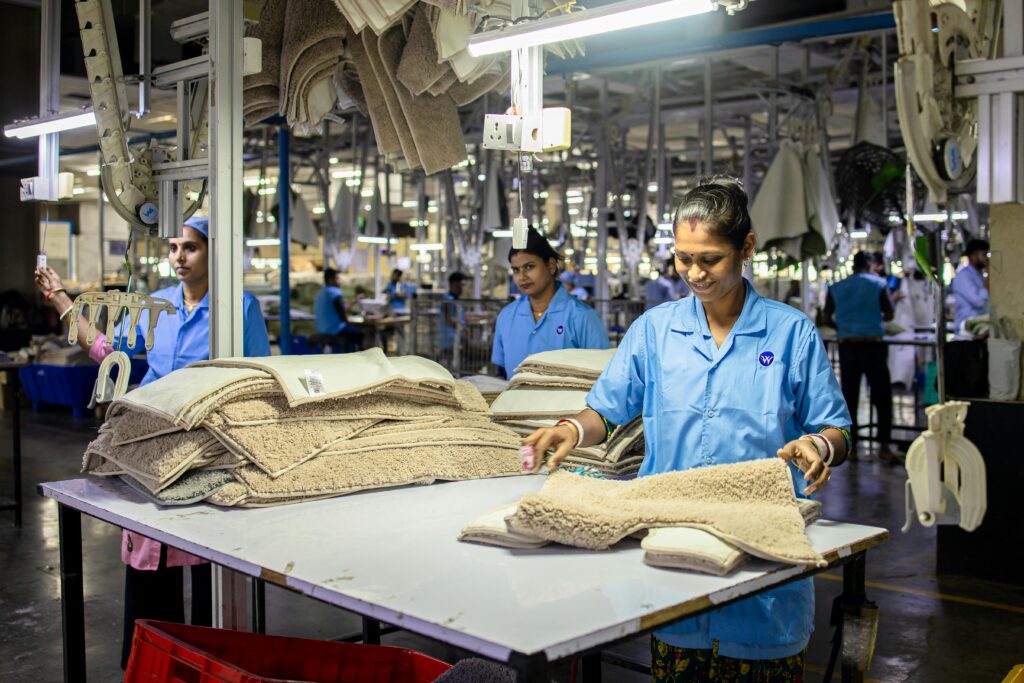 pexels-photo-32399708-32399708 Smiling textile factory workers sorting beige carpet mats in a well-lit industrial setting.